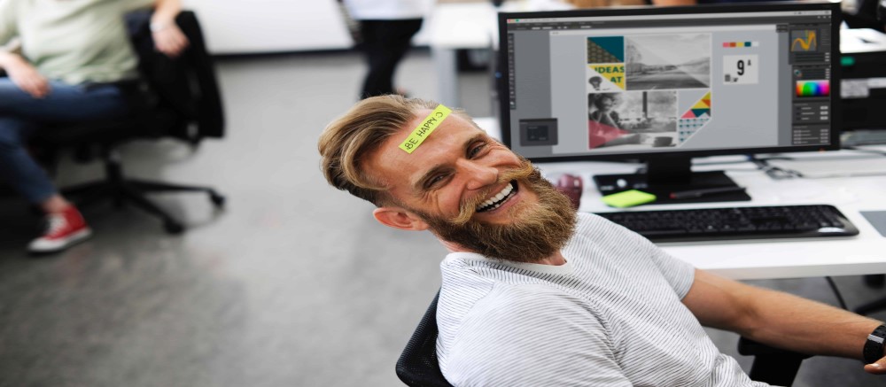 Bearded man sitting at work laughing, enjoying his occupation, while having a sticker attached to his forehead